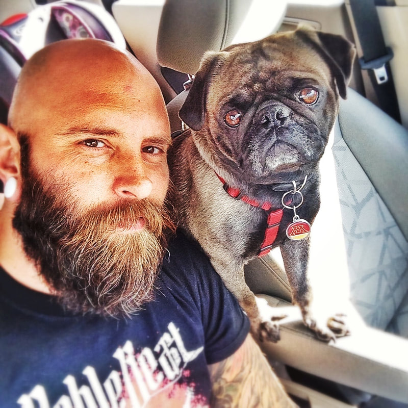 Slight side profile of male, head shaved with brown beard in a car with a pug, both looking into the camera.