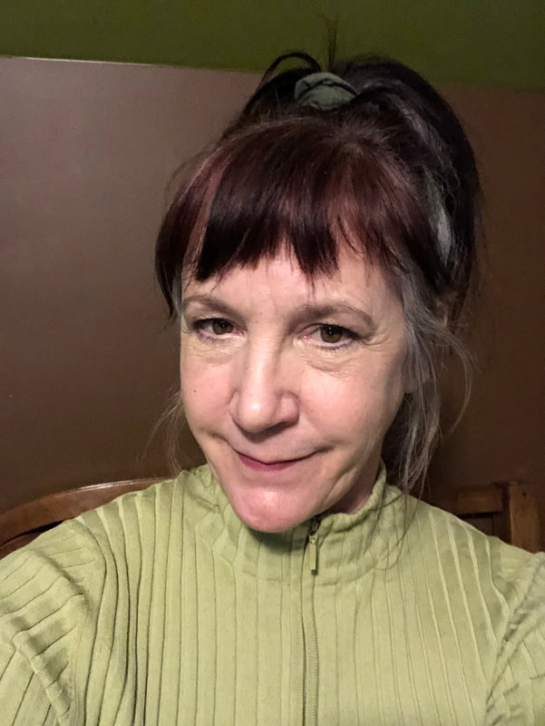 Photo of a woman. She is Caucasian and a “brunette “ with bangs. Her hair is pulled up high on her head in a bun. She has brown eyes and minimal makeup. She is wearing an avocado colored zip up turtleneck sweater. Background is a wall. It is mostly painted chocolate brown. The upper portion is painted olive green.