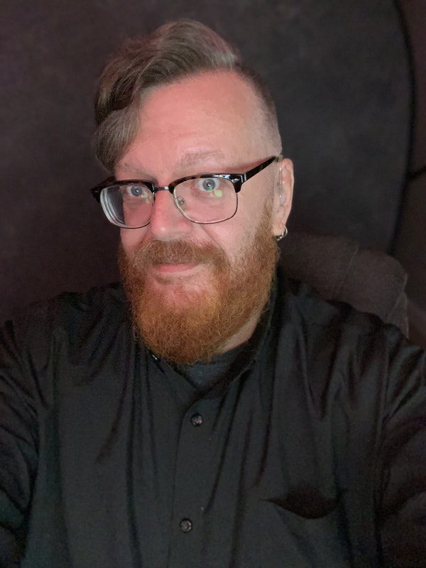 White cis male with light brown hair and some gray strands, blue eyed, retro eyeglasses, small silver hoop earrings and coiffed red beard wearing black shirt with black background.