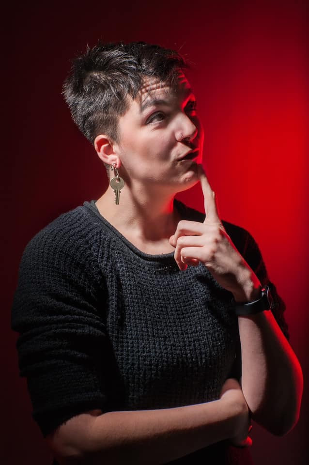 Side view of Jess (they/them) wearing a black shirt, sporting a key earring and an inquisitive smirk with their pointer finger under their chin, in front of a red backdrop. Their eyes are looking up as if they are thinking “whodunnit?”, as this was a promo picture from a play they were recently involved with.