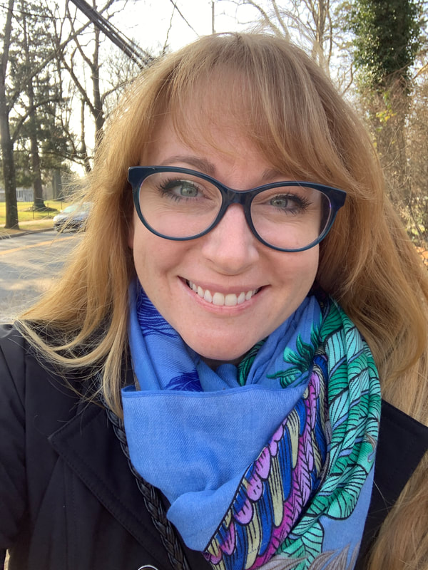 White female with strawberry blonde hair and green eyes smiling directly into the camera. She is wearing large blue glasses, a blue scarf with multi-colored feathers, and a black jacket. Photo taken outdoors with a street and trees in the background.