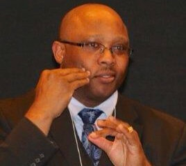 African-American gentleman with almost completely bald, brown eye with black semi-rimless frame include non-reflective lens, medium build wearing a light blue formal shirt and paisley pattern blue and light blue tie with full black suit.  My hand is sign as “home” on my right face slightly below my cheek while giving a short comment at the Award ceremony.