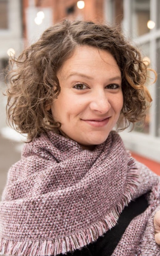A curly-haired white woman is smiling. She is wearing a black shirt and has a pink scarf around her neck.