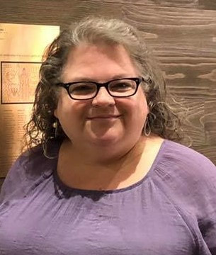 Facing the camera, Amy, white lady, wearing light purple tunic with black rectangle framed eyeglasses with mixture of gray and light brown hair in it's own curls.  The background is several wood shiplaps. On the left there's a gold plague with wordings that reflected the Signing Starbucks in Washington D.C.