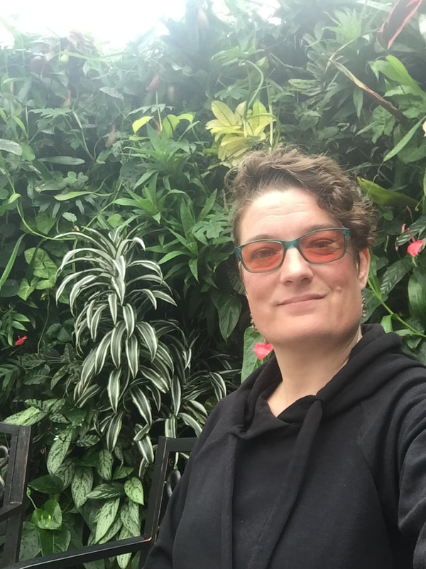 Jennifer, white woman with short curls and an undercut, wears pink tinted glasses and a black top, smiling into the camera in front of a wall of green leafy plants.