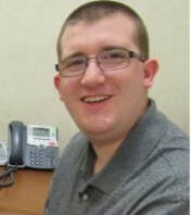 A white male with glasses, with short brown hair in a buzz cut. He is wearing a gray-collared shirt sitting next to a desk phone and smiling into the camera. 