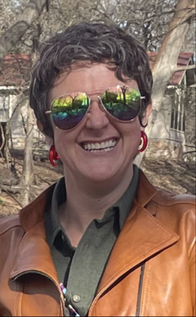 Pavey, a  white female with short salt and pepper hair wearing a golden rainbow mirrored aviator sunglasses, Forest green button up collared shirt with  a dark camel colored leather jacket, and is smiling at the camera. Trees in the background. 