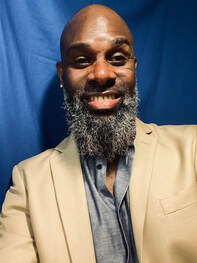 Koko, a Black DeafBlind man, with a bald head and a long salt and pepper curly beard, is wearing a gray button up shirt and cream colored suit jacket, and is looking directly into the camera and smiling.  The background is dark blue. 