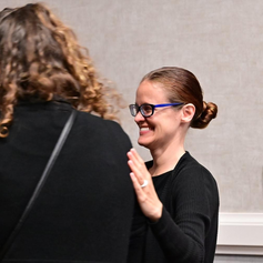 Krystal Sanders, a white woman with brown hair pulled back in a bun. She has black and blue glasses. She is wearing a long-sleeved black sweater. She smiles widely as her hand is on Jelica’s back, which is turned from the camera. Jelica is conversing with Heather Holmes in Protactile; Heather is seen laughing. (If can be seen in the cropped photo). They are in a meeting room with light grey walls and white trimming.