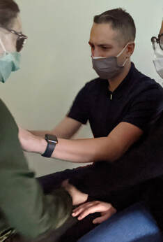 A white male wearing a zip-up polo shirt is fully concentrated as he interpretes into protactile on the chest of a person seated across from him. A third person is in contact and watches the interaction. All three are wearing facemasks.  