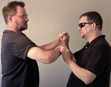 Jaz (right), a Caucasian DeafBlind man with short hair, wearing sunglasses and a dark shirt, using Protactile language with another person.