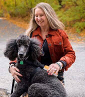 A white female with long blonde hair blowing in the wind, wearing an orange colored jacket sitting on the side of a road full of trees.  She is looking down and smiling at her black poodle dog. Her right hand is on his chest and left hand over his back as he lies on her lap and looks directly into the camera. 