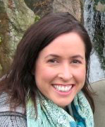 Close up photo of on person, Erin Frost. She is a female, caucasian, in her early 40s, with brown eyes, and shoulder-length dark brown hair, parted on the side. She is standing in front of a rock waterfall, wearing a green and white scarf around her neck and smiling at the camera.