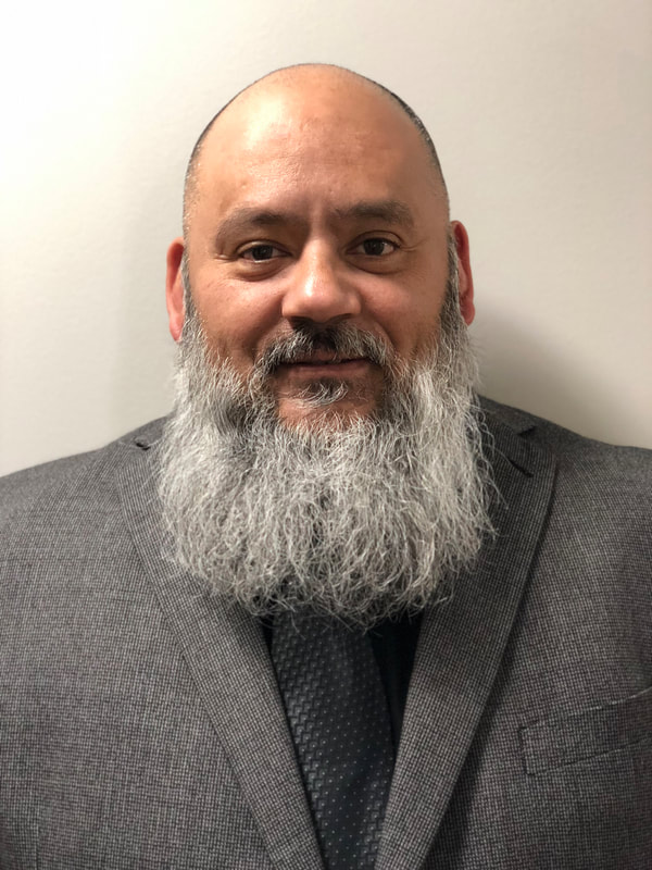 Picture of an older Latinx male with a long mostly white beard wearing a gray sports coat and tie.  white wall background
