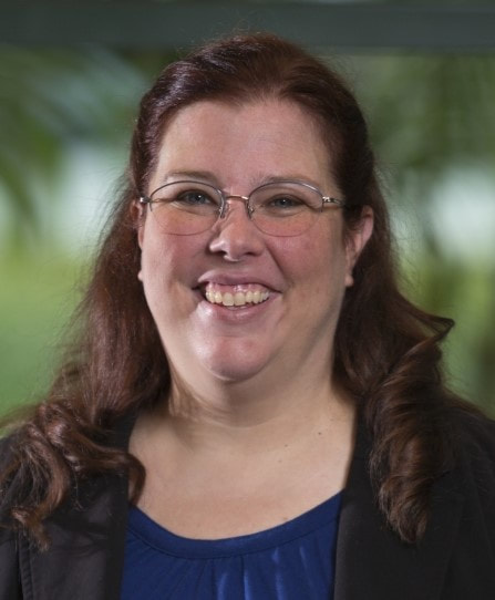 Image Description: Head shot from just below the shoulders of a smiling, heavyset white woman with blue eyes and shoulder-length auburn hair wearing oval, wire-rimmed glasses, a dark blue scoop-necked top and a black blazer in front of a blurred background of greenery with a white building.