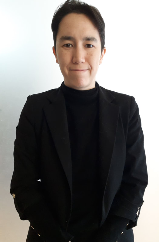 38-year-old nonbinary person with light skin, brown eyes, and short black hair, small and thin, wearing a black shirt and jacket. The person is bi-racial (Asian and white). The photo shows their face and torso and has a light gray background. Pronouns: they/them.