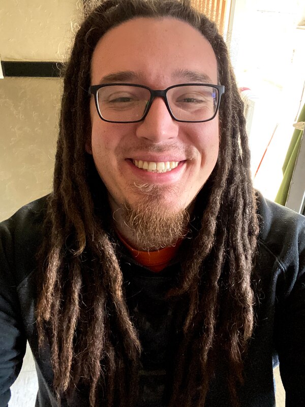 "He/Him/His
Hispanic/Latinx

A light-skinned young man smiles at the camera.  He has long brown dreadlocks that reach halfway down his chest.  He has square-shaped, black-framed glasses and a reddish-brown goatee that reaches 1 inch off of his chin.  There is some stubble of facial hair around his upper lip and cheeks.  He is wearing a black/grey sweater with an orange shirt underneath.  Behind him is an open door and a tan/beige wall with black trim."