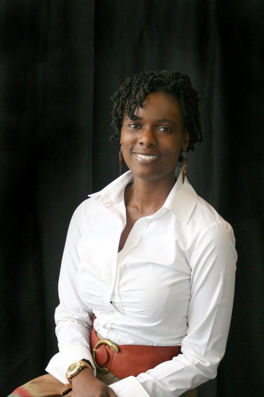 An African-American woman (brown eyes, black hair in two-strand twists) sits smiling,  in a white dress shirt against a black background.