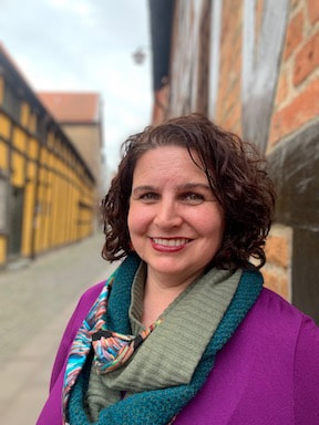 A smiling white woman (her, hers, her) with short brown curly hair and brown eyes standing in an old European street lined with old brick and wooden buildings.