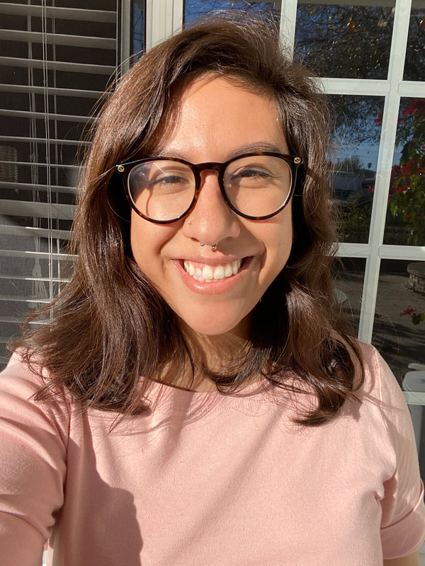 Tricia Vazquez, a Brown-Skinned (skin color like adding medium amount of creamer to your coffee) Chicana taking a selfie. She is outside standing in front of an open window. There's blinds behind her and a reflection of the front yard in the window. The sunlight is hitting her face. Her hair length is touching her shoulders and the color is a rich brown. She has black round glasses on, dark brown eyes, and squinting.  Her nose septum is slightly crooked. She's smiling at the camera.