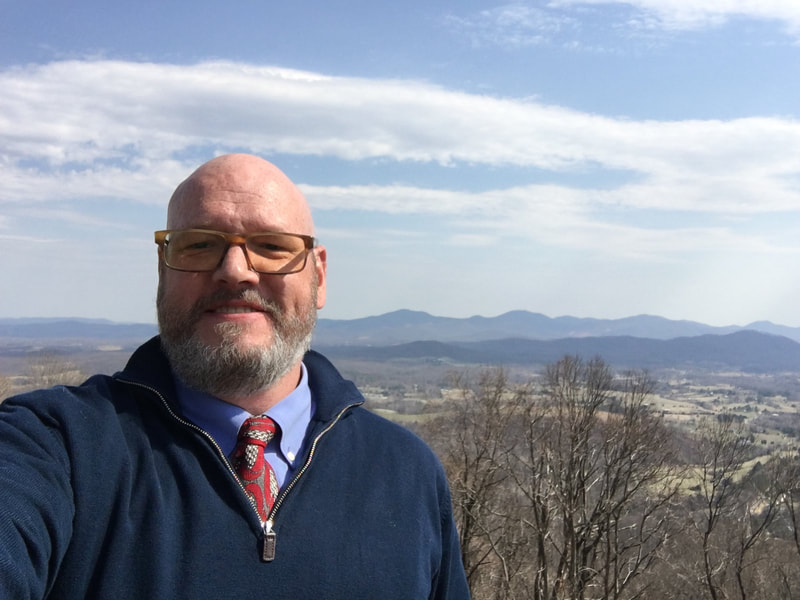 Smiling white bald male with a short salt and pepper beard and brown glasses wearing a navy blue zipped sweater over a blue collared shirt and patterned red tie with bare trees, mountains, and clouds in the background