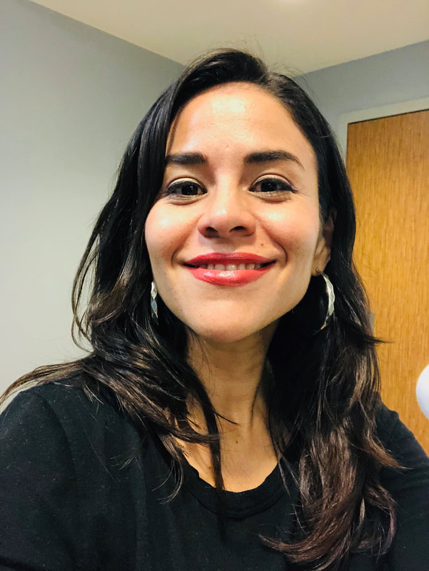 [ Background: grey wall and wooden door. Latin woman smiling wearing red libpstick. Dark  hair a few inches below shoulder level. Dark eyes. She is wearing large silver ring earings and a black shirt]