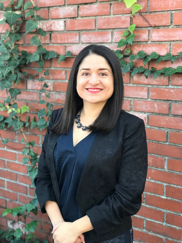 Irene Robles, female Mexican-American, fair skin, brown eyes, dark brown hair, wearing navy shirt with black blazer and navy necklace. Background is red brick wall with vines hanging down from it.