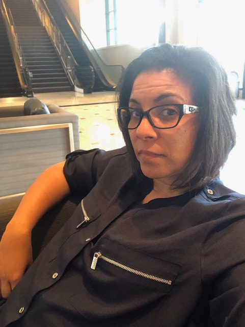 Selfie photo of biracial Amalia sitting in the chair with right arm on the armrest in dark blue blouse with silver buttons/zippers and brown hair and escalators and stairs in the background.