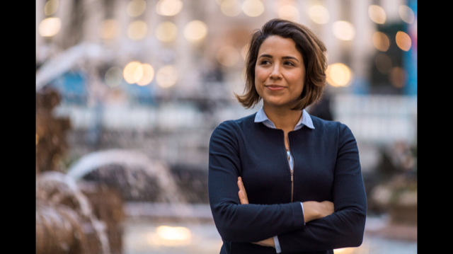In the photo is Latina woman standing with her arms crossed and bright smile on her face looking up towards the future. She is wearing a light blue collard shirt with a dark blue blazer. The background is faded out with a fountain and water shooting out and lots of white lights.