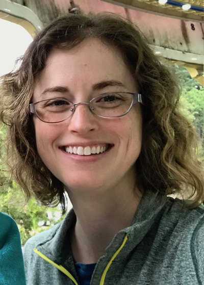 White woman smiling directly at the camera. She has curly brown shoulder-length hair, glasses, and freckles. She is wearing a light green zip-up jacket with a yellow zipper. In the back ground is the white and red faded wooden overhang of the carousel at the Oregon Zoo.