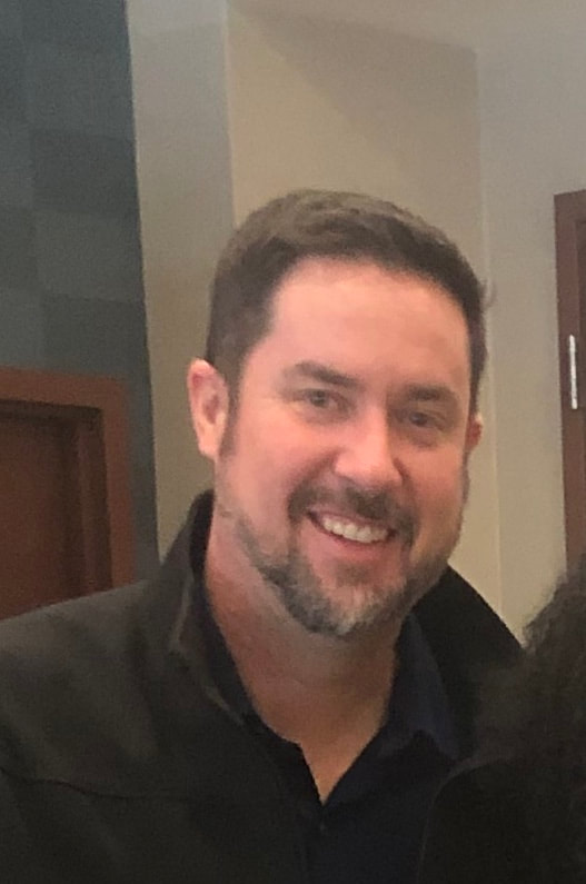 Smiling white male with dark short hair, blue eyes, trimmed gray and black beard, wearing a black shirt and black coat