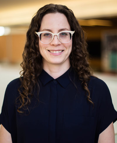 A Caucasian woman in her late 30's with long curly brown hair and blue-green eyes smiles at the camera.  She is wearing a pair of pearl-rimmed eyeglasses and short-sleeved navy blue blouse.