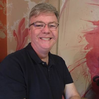 A Caucasian man in his 50's with short cut, slightly greying hair. He is smiling and has glasses. He is wearing a black short-sleeved shirt and sitting in front of a red and tan colored wall.