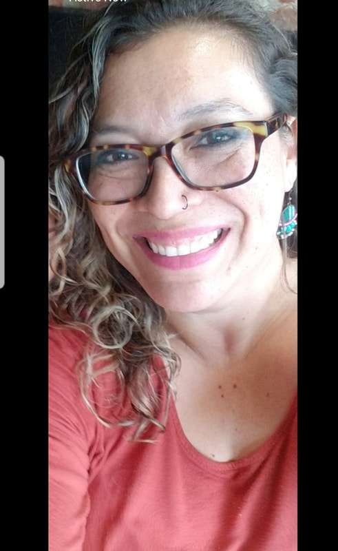 A light skinned Latina with brown and blond curly hair. she is smiling at camera and is wearing eye make up, brown glasses, a black nose ring, and magenta lipstick. She is wearing a salmon color shirt and an earrings are a mix of colored beads in turquoise, white, and black, though you can only see one.