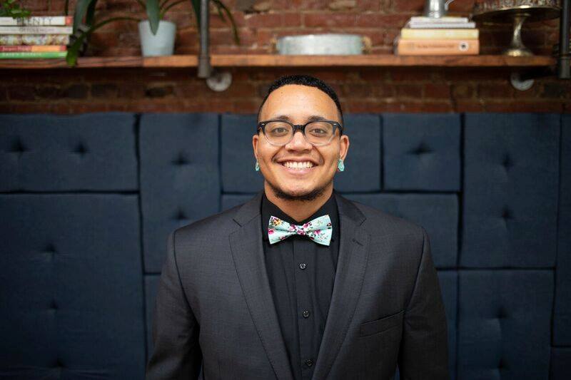 MJ, a Black/Filipinx and light brown skin Trans masculine person, is standing in front of a brick wall with a shelf of potted plants and stacks of books on it. On the bottom half of the wall is navy colored fabric backing to a restaurant booth. MJ is the focal point of the image and is smiling toward the camera wearing their black rimmed glasses, a big smile, turquoise gauges in their ears, and a dark grey suit with a black button-up shirt. MJ has dark brown eyes, short black hair and a short beard beneath their chin. MJ is also wearing a turquoise/light blue bow tie that has floral print.