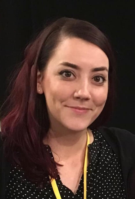 A red haired, brown eyed, white woman is wearing a black v-neck blouse with small white polka dots and a solid black sweater over her shoulders..  She is smiling at the camera while standing behind a solid black backdrop, with a yellow lanyard around her neck.