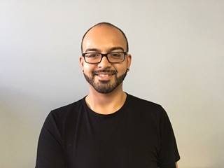 Latinx male, bald with brown eyes and trimmed beard and black framed glasses and black shirt smiling and looking towards the camera.