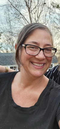 White female with brown hair in a ponytail with bangs on one side.  She is  wearing brown and black frame eyeglasses with a black scoop t shirt and smiling at the camera. 