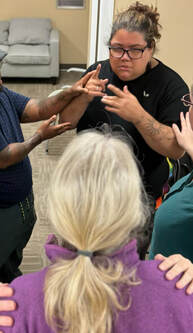 Yash, a DeafBlind Latine Queer woman, showing sun shape tattoo on her left arm wearing a black shirt, black rimmed glasses and black earrings with dark brown and dirty blonde hair pulled back in a bun using Protactile sitting in a group. Each person in the group has their hand on each other.