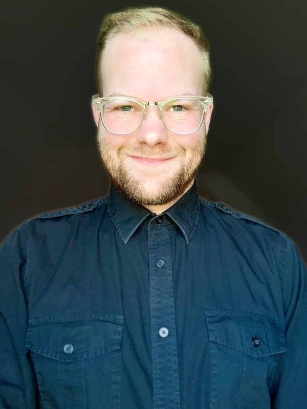 A white masculine person looking directly ahead, a neutral smile. They are blond with blue eyes, has a short beard of varying brown and blond colors, have glasses that are clear on the top and silver rims on the bottom, and is wearing a black button shirt. Behind him, there are oddly angled walls that are a white color, but they are completely plain with no decoration.