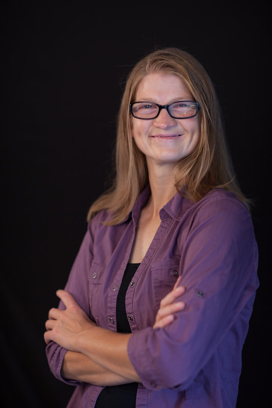 A white woman with dirty blonde, shoulder length hair, wearing black frame glasses, with dimples. Wearing  a purple blouse with a black t-shirt underneath. Her arms are folded and she is standing in front of a black background.