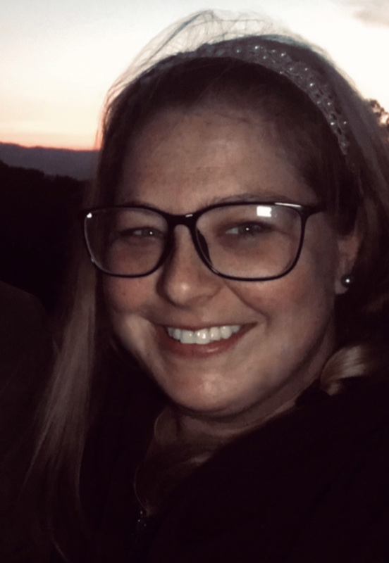 Feminine presenting smiling white woman with blue eyes behind large black rimmed glasses is looking straight into the camera. Her long blonde hair is held back by a tan and pearl headband with some strands flying in the wind. The background is peach-orange with black hills, indicating a sunset.