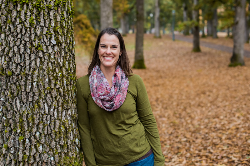 A woman (Susie) in a park during Fall.  Susie is wearing a green long sleeve shirt with a multi-colored scarf of blue, burgundy and white, jeans and is smiling.  Susie is leaning against a tree trunk that has some green moss on it. In the background there are fallen yellow leaves on the ground as well as a path and other trees.
