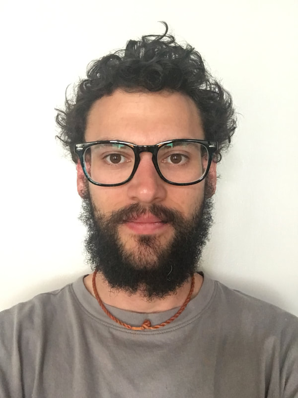 Close up portrait of Tullos' face in white background, a man showing slightly smirks with dark curly brown hair and beard, brown eyes with black-ish glasses frame, he is wearing gray t-shirt with orange necklace.