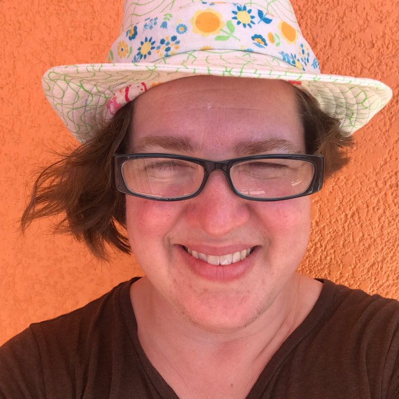 white woman with a white and green fedora with a floral band. Wearing black glasses and an uneven brownish hair cut that comes to her lips on one side and her eyes on the other. Wearing a brown v-neck shirt and standing against an orange stucco wall. She is smiling and squinting into the sun with rosy cheeks.