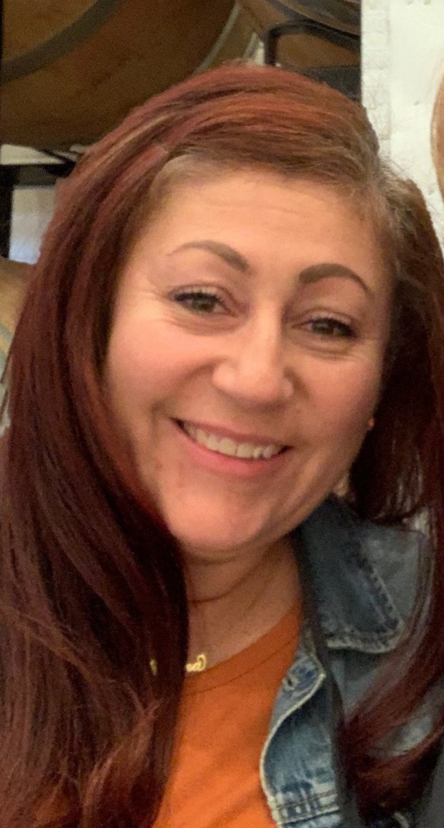 White female with red/brown long hair, rusty orange shirt with denim jacket, and wearing a gold necklace "Malena". Her face is big smile.
